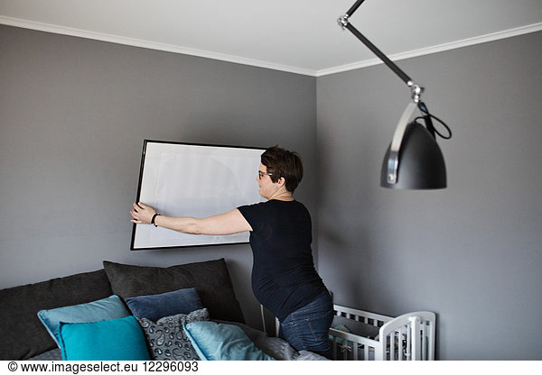 Pregnant female arranging paintings on wall in bedroom at home