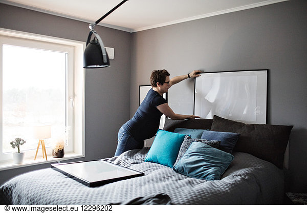 Pregnant woman arranging paintings on gray wall in bedroom at home