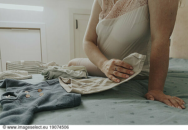 Pregnant woman choosing baby clothing on bed at home
