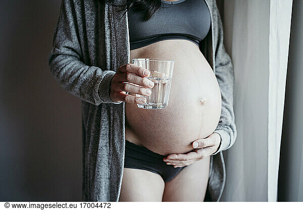 Pregnant woman holding drinking glass while touching belly at home