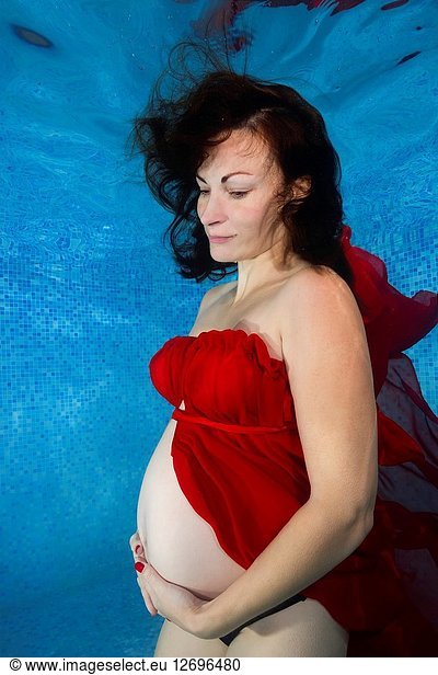 Pregnant woman in a red dress posing underwater in the pool.