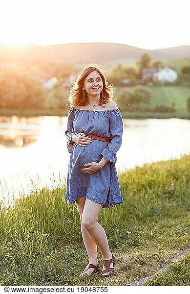 pregnant woman in blue dress holds her belly under rays of sun
