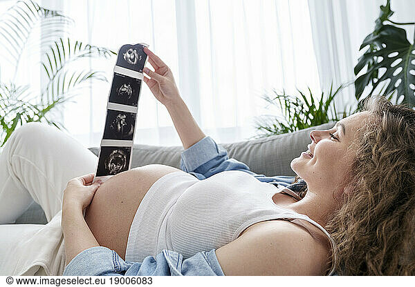 Pregnant woman looking at ultrasound scans lying on couch at home