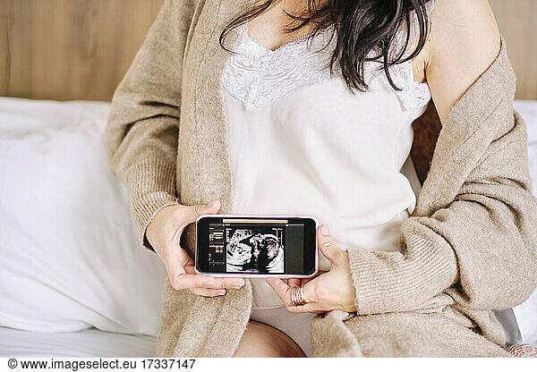Pregnant woman showing ultrasound image on smart phone at home