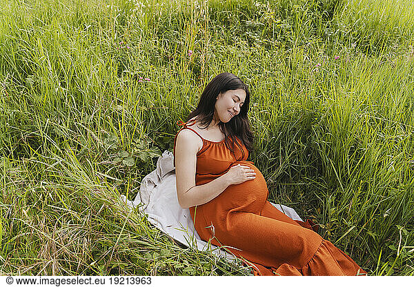 Pregnant woman sitting on blanket near grass in meadow