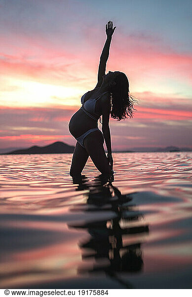 Pregnant woman stretching arm standing in sea water at sunset