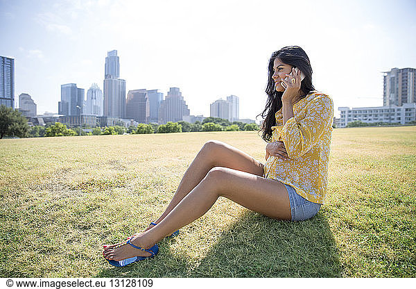 Pregnant woman using phone while sitting on grass against city