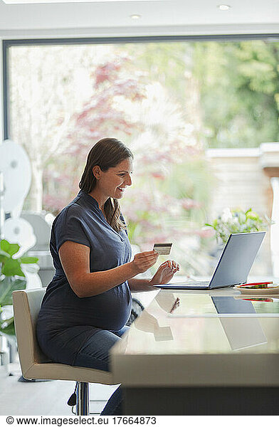 Pregnant woman with credit card online shopping at laptop in kitchen