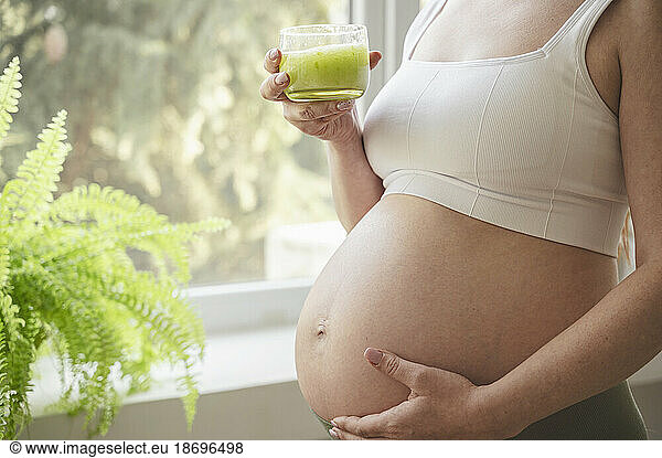 Pregnant woman with hand on stomach holding glass of green smoothie at home
