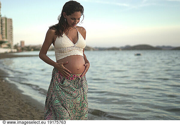 Pregnant woman with hands on stomach standing at beach