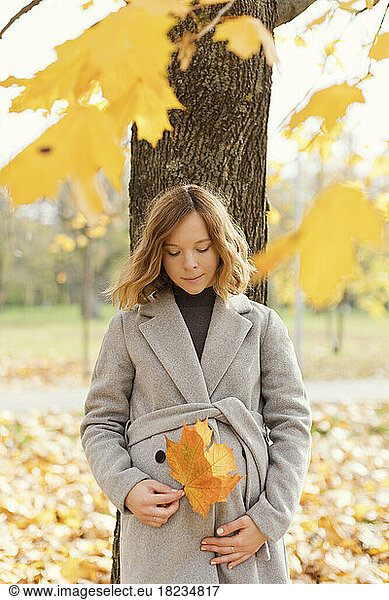 Pregnant woman with maple leaf leaning on tree at autumn park