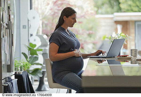 Pregnant woman working from home at laptop in kitchen