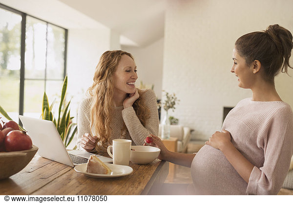 Pregnant women eating cake and fruit at laptop in dining room