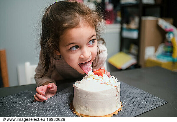 Preschool age girl licking birthday cake and looking off camera