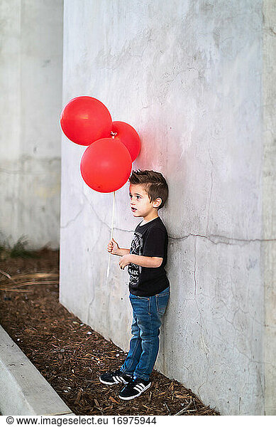 Preschooler boy leaning against concrete wall holding red balloons.