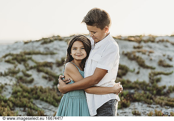 Preteen boy in white shirt hugging his younger sister at the beach