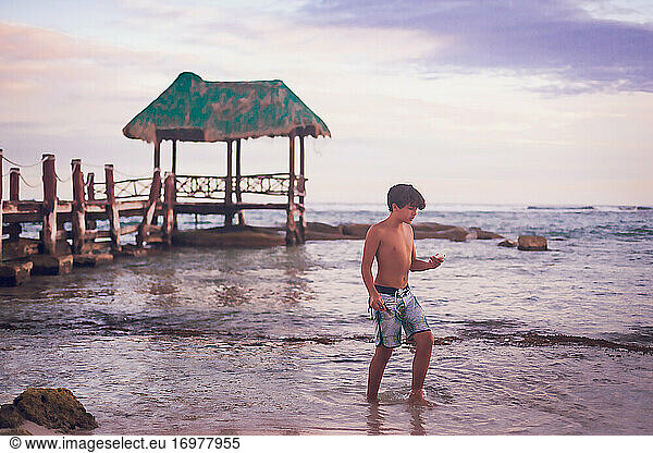 Preteen boy walking on the beach with a wooden pier in the background.