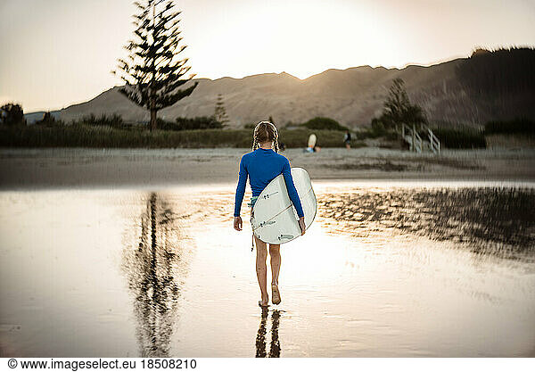 Preteen girl on beach at dusk carrying surfboard