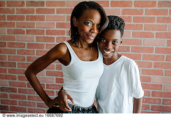 Pretty black mom hugging smiling preteen son in front of brick wall