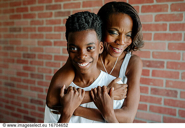 Pretty black mom hugging smiling preteen son in front of brick wall