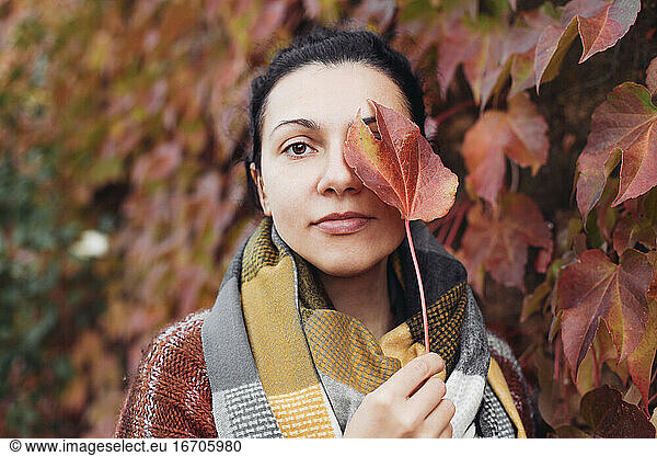 Pretty woman covers her face with a red leaf and looking at the camera