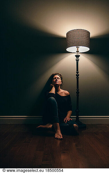 Pretty woman sitting alone on the floor beside a lamp in dark room.
