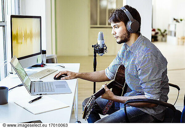 Professional with headphones and guitar using laptop while sitting at recording studio