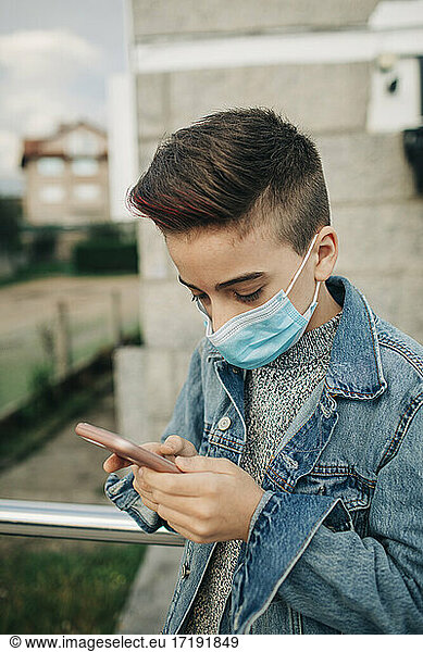 Profile teen boy wearing mask and denim jacket using cellphone outdoor