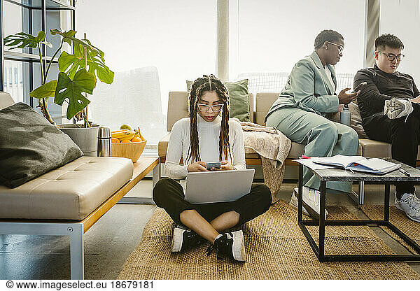 Programmer using mobile phone while sitting with laptop on carpet in creative office