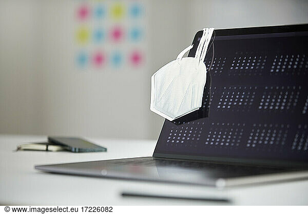 Protective face mask hanging on laptop screen at desk in office
