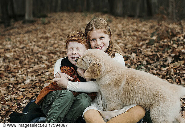 Puppy licking boy's face sitting with sister on autumn leaves in forest