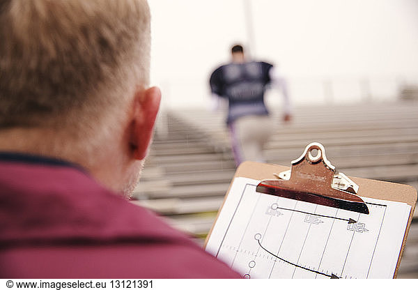 Rückansicht des Trainers  der die Zwischenablage hält  mit Blick auf den im Stadion trainierenden American-Football-Spieler