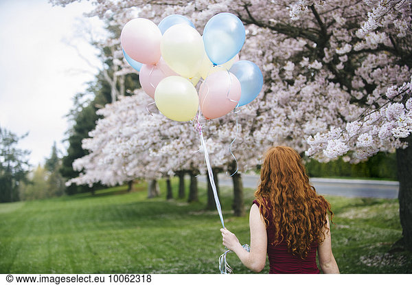 Rückansicht einer jungen Frau mit langen  gewellten roten Haaren und Luftballons im Frühjahrspark