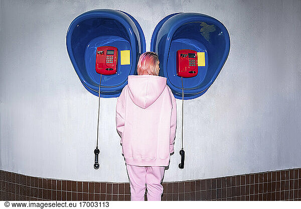 Rückansicht einer jungen Frau mit rosa Haaren und rosa Kapuzenshirt  die vor einer Telefonzelle steht