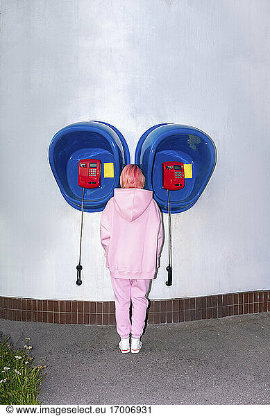 Rückansicht einer jungen Frau mit rosa Haaren und rosa Kapuzenshirt  die vor einer Telefonzelle steht