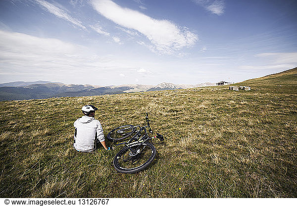 Rückansicht eines auf Grasfeld sitzenden Mountainbikers gegen den Himmel