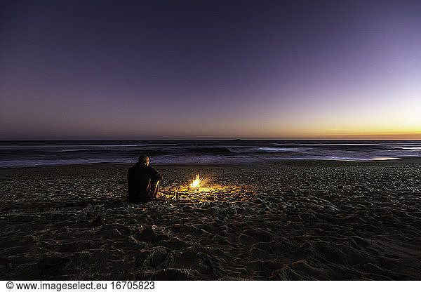 Rückansicht eines einsamen Mannes und Lagerfeuer am Strand bei Sonnenuntergang