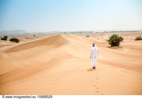 Rückansicht eines jungen Mannes aus dem Nahen Osten in traditioneller Kleidung  der in der Wüste spazieren geht  Dubai  Vereinigte Arabische Emirate