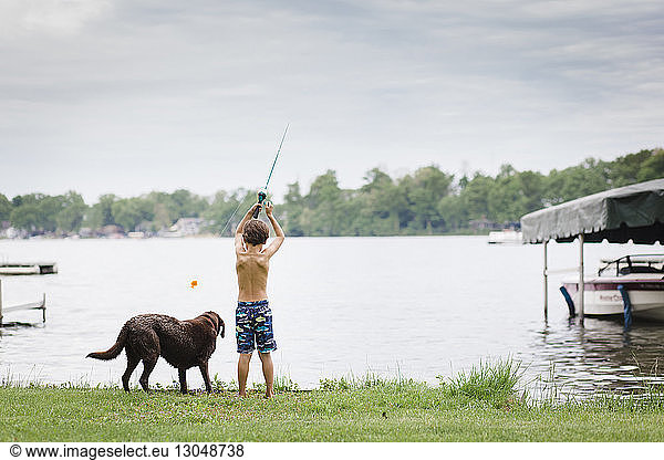 Rückansicht eines Jungen ohne Hemd beim Angeln  während er mit seinem Hund am Seeufer vor bewölktem Himmel steht
