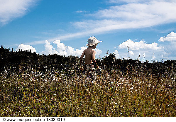 Rückansicht eines Jungen ohne Hemd mit Hut  der auf dem Feld vor bewölktem Himmel steht