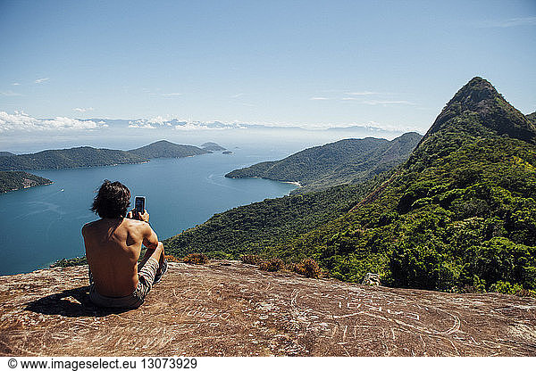 Rückansicht eines Mannes  der das Meer fotografiert  während er auf einem Berg gegen den Himmel sitzt
