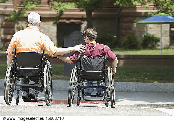 Rückansicht eines Mannes mittleren Alters und einer Frau mittleren Alters mit Muskeldystrophie  die in Rollstühlen eine Straße überqueren