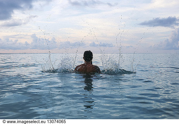 Rückansicht eines Mannes ohne Hemd  der Wasser im Meer gegen bewölkten Himmel spritzt