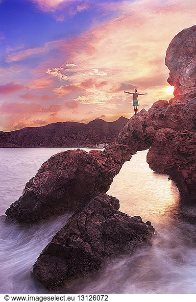 Rückansicht eines Mannes ohne Hemd mit ausgestreckten Armen  der bei Sonnenuntergang auf einem Felsen über dem Meer gegen den Himmel steht