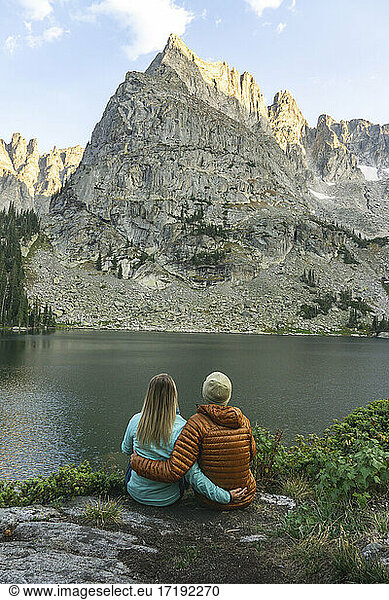 Rückansicht eines Paares mit Blick auf den Lone Eagle Peak  während es mit ausgebreiteten Armen am Seeufer sitzt