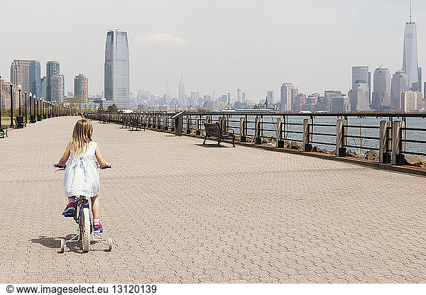 Rückansicht eines radfahrenden Mädchens auf der Promenade mit der Skyline der Stadt im Hintergrund