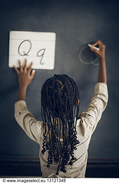 Rückansicht eines Schulmädchens mit geflochtenem Haar  das das Alphabet Q an die Tafel schreibt