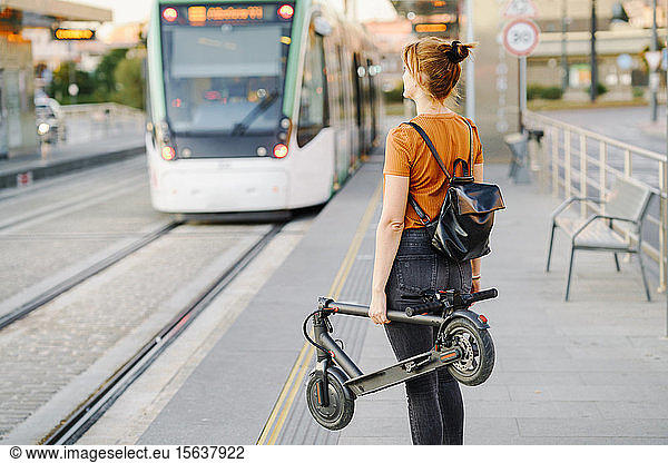 Rückenansicht einer Frau mit Rucksack und gefaltetem Elektroroller  die an der Straßenbahnhaltestelle wartet