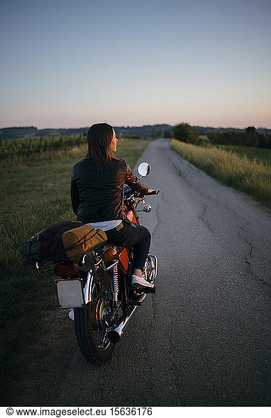 Rückenansicht einer jungen Frau  die bei Sonnenuntergang auf einem Motorrad sitzt und auf die Aussicht schaut  Toskana  Italien