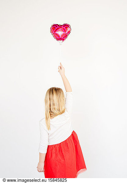 Rückenansicht kleines blondes Mädchen in rotem Tutu  das einen roten Herzballon hält